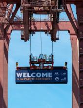 A shipping container with banner that reads “Welcome to the Pacific Northwest” hangs from a crane at Terminal 46 in Seattle.
