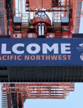 A shipping container with a banner that reads “Welcome to the Pacific Northwest” hangs from a crane at Terminal 46 in Seattle.