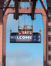 A shipping container with a banner that reads “Welcome to the Pacific Northwest” hangs from a crane at Terminal 46 in Seattle.